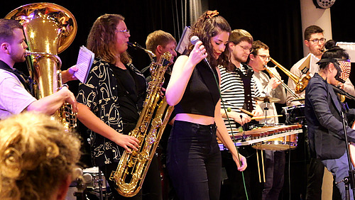 Several people playing wind and percussion instruments, along with one person holding a microphone, performing on stage