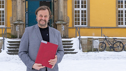  Prof. Focks steht mit Ernennungsurkunde in der Hand im verschneiten Schloss-Innenhof und l&auml;chelt in die Kamera