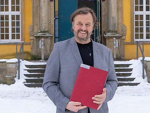 Prof. Focks steht mit Ernennungsurkunde in der Hand im verschneiten Schloss-Innenhof und l&auml;chelt in die Kamera