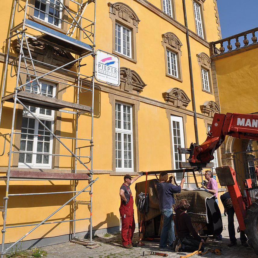 Blick auf die Aussenfassade des Osnabrücker Schlosses. Im zweiten Stock ist ein Fenster geöffnet, davor ist ein Gerüst aufgebaut. Unten steht ein Flügel auf der Seite auf dem Gestell eines Krans. Drei Personen befestigen Transportbänder an dem Instrument.