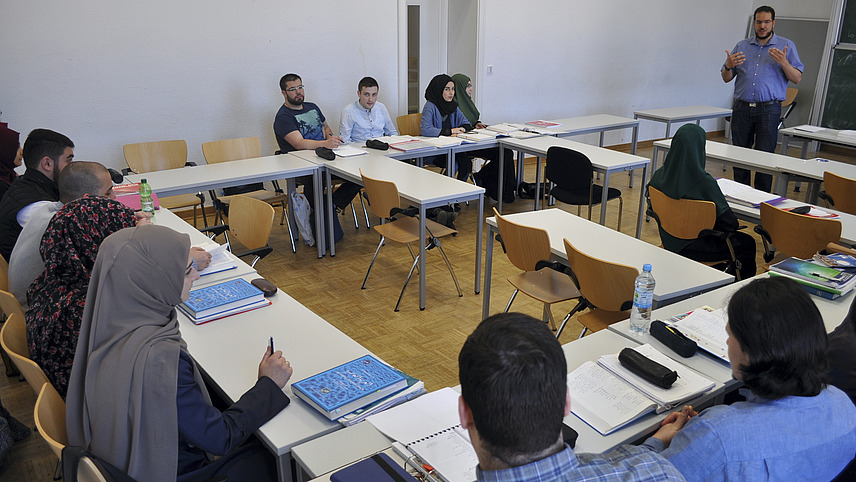 Female students with hijab and male students sit in a seminar room with work materials and listen to the lecturer.