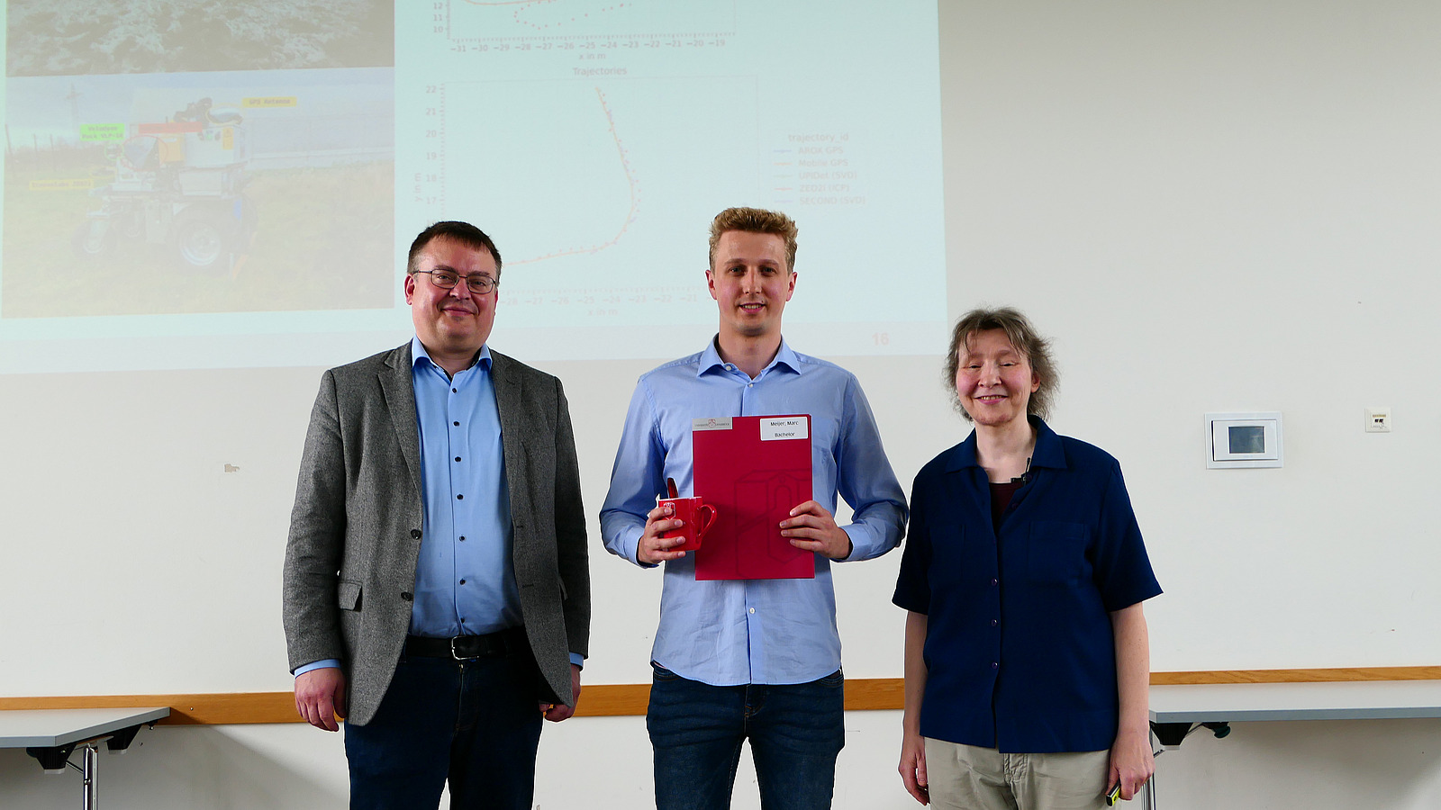 Ein Student mit Studiendekanin und Dekan stehen nebeneinander. Der Student hat eine Mappe in der Hand