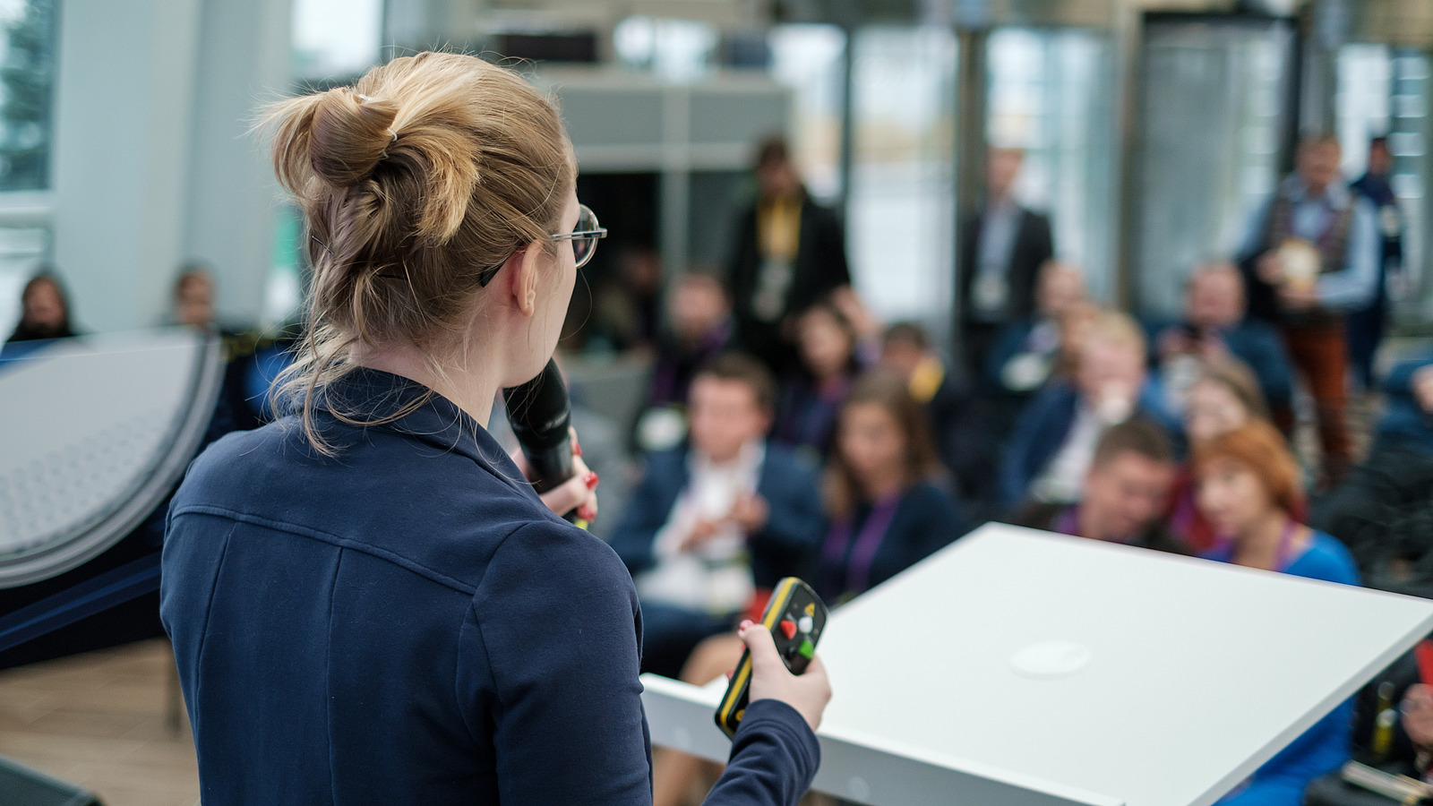 A speaker from behind with remote control and microphone, audience in the background