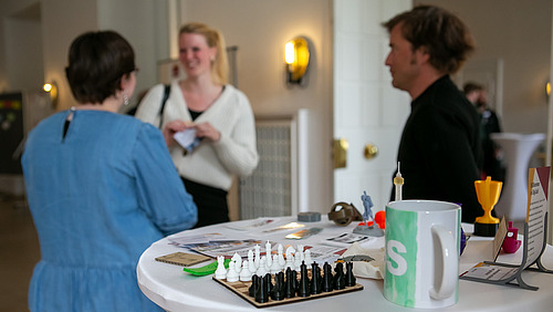 Drei Personen stehen bei einem Stehtisch und unterhalten sich. Im Vordergrund sieht man auf dem Tisch ein kleines Schachspiel und einen Becher