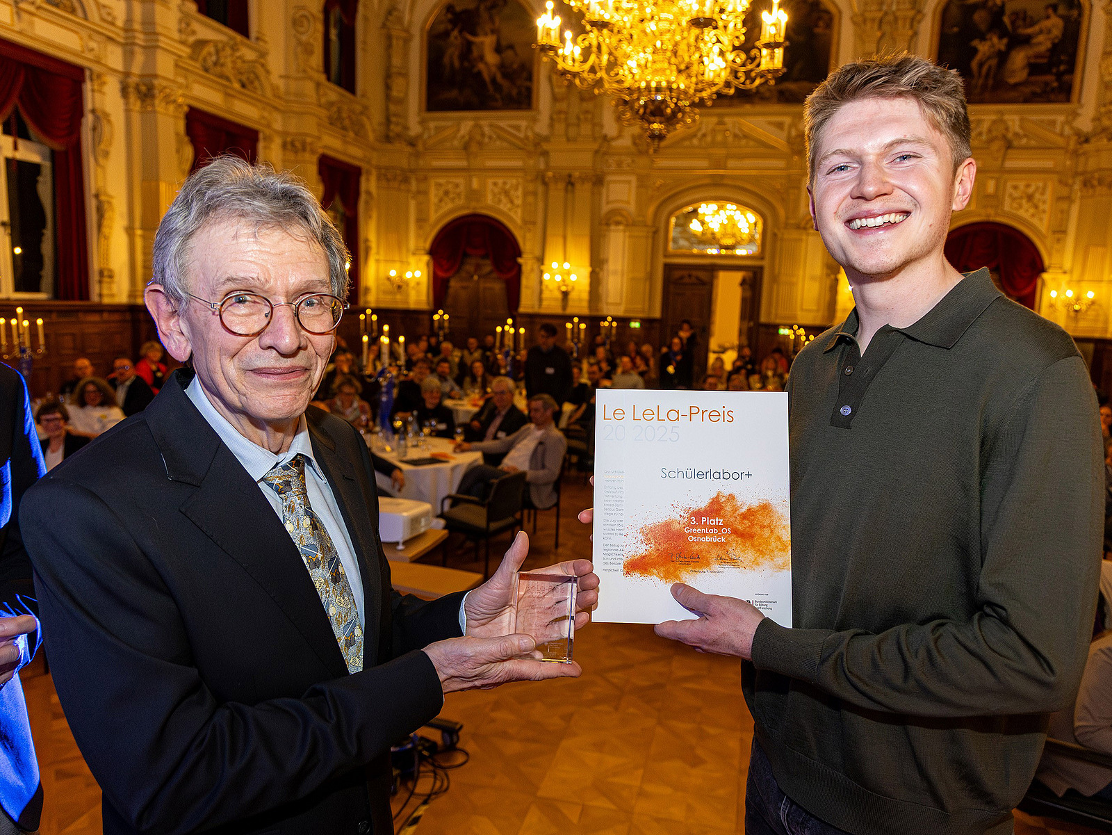 Zwei Personen halten einen Preis und eine Urkunde in die Kamera. Im Hintergrund sitzen in einem Saal viele Personen.