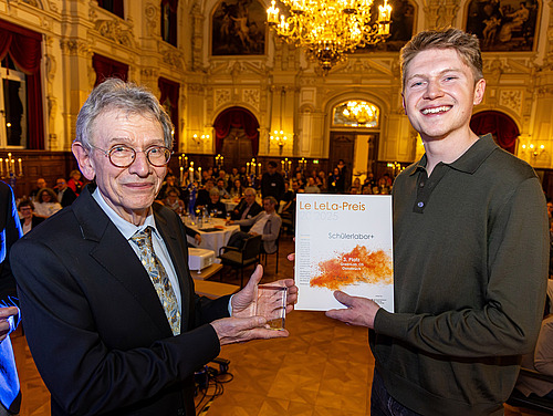 Zwei Personen halten einen Preis und eine Urkunde in die Kamera. Im Hintergrund sitzen in einem Saal viele Personen.