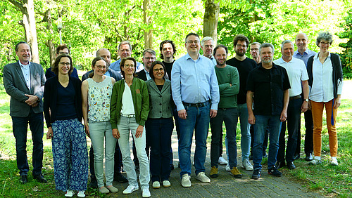 Etwa 20 Personen stehen unter Bäumen auf einem Weg zwieschen zwei Grasflächen.