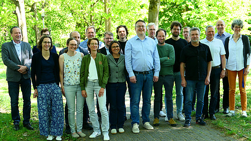 About 20 people are standing under trees on a path between two lawns.