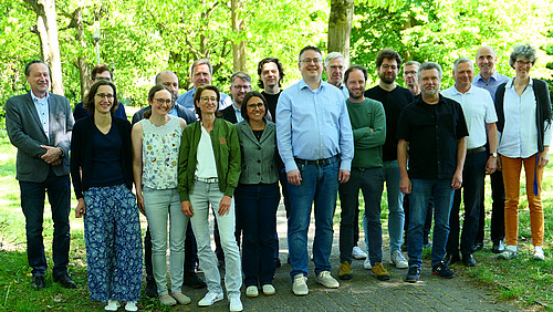 Etwa 20 Personen stehen unter Bäumen auf einem Weg zwieschen zwei Grasflächen.