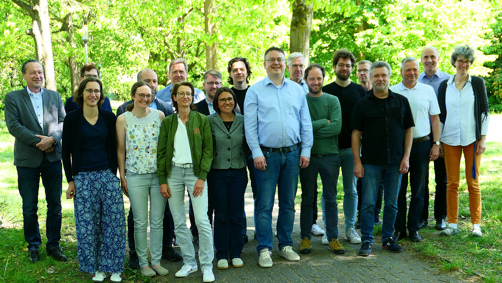 Etwa 20 Personen stehen unter Bäumen auf einem Weg zwieschen zwei Grasflächen.