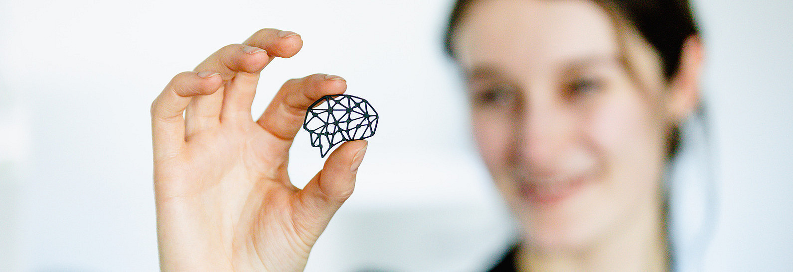 Eine Person hält mit Daumen und Zeigefinger ein kleines, schwarzes, geometrisch gefertigtes Objekt, das die Form eines Gehirns hat. Im Hintergrund ist die Person unscharf zu erkennen. Sie lächelt und schaut auf das geometrische Objekt. Die Szene ist hell beleuchtet und der Hintergrund wirkt einfach und minimalistisch.