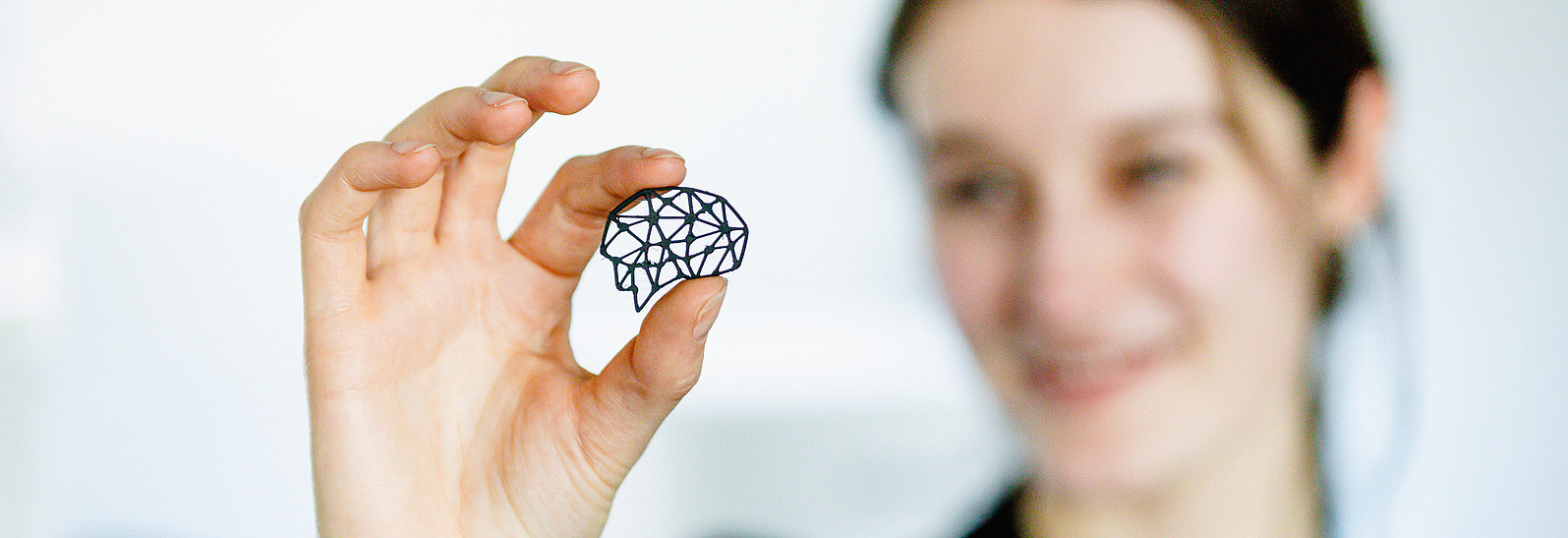 A person is holding a small, black, geometrically shaped object in the form of a brain with their thumb and index finger. The person is blurred in the background. She is smiling and looking at the geometric object. The scene is brightly lit and the background appears simple and minimalist.