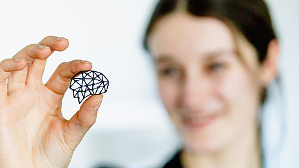 Eine Person hält mit Daumen und Zeigefinger ein kleines, schwarzes, geometrisch gefertigtes Objekt, das die Form eines Gehirns hat. Im Hintergrund ist die Person unscharf zu erkennen. Sie lächelt und schaut auf das geometrische Objekt. Die Szene ist hell beleuchtet und der Hintergrund wirkt einfach und minimalistisch.