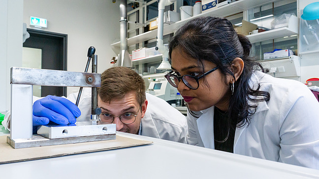 Eine junge Frau und ein junger Mann in wei&szlig;en Laborkitteln blicken konzentriert auf eine experimentelle Apparatur. 