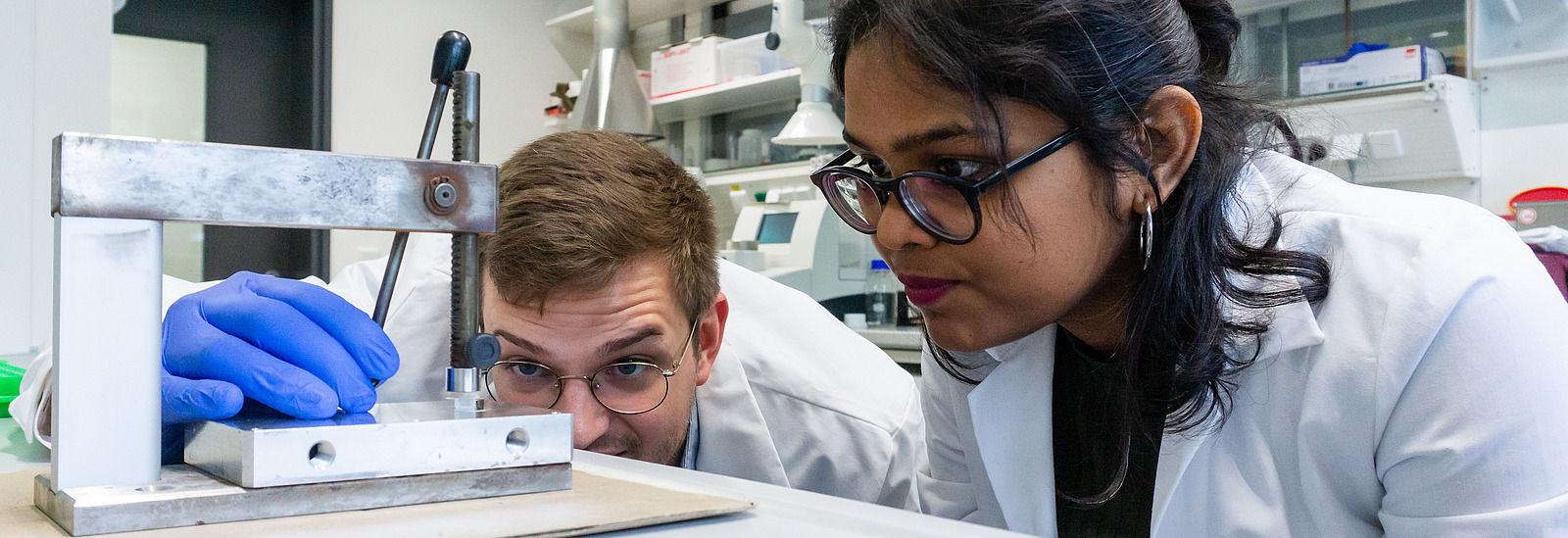 Eine junge Frau und ein junger Mann in wei&szlig;en Laborkitteln blicken konzentriert auf eine experimentelle Apparatur. 