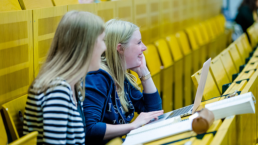 Zwei lächelnde Studentinnen sitzen in einer Vorlesung auf gelben Sitzbänken. Vor ihnen liegen Schreibutensilien, ein aufgeklappter Laptop und ein Gesetzbuch.