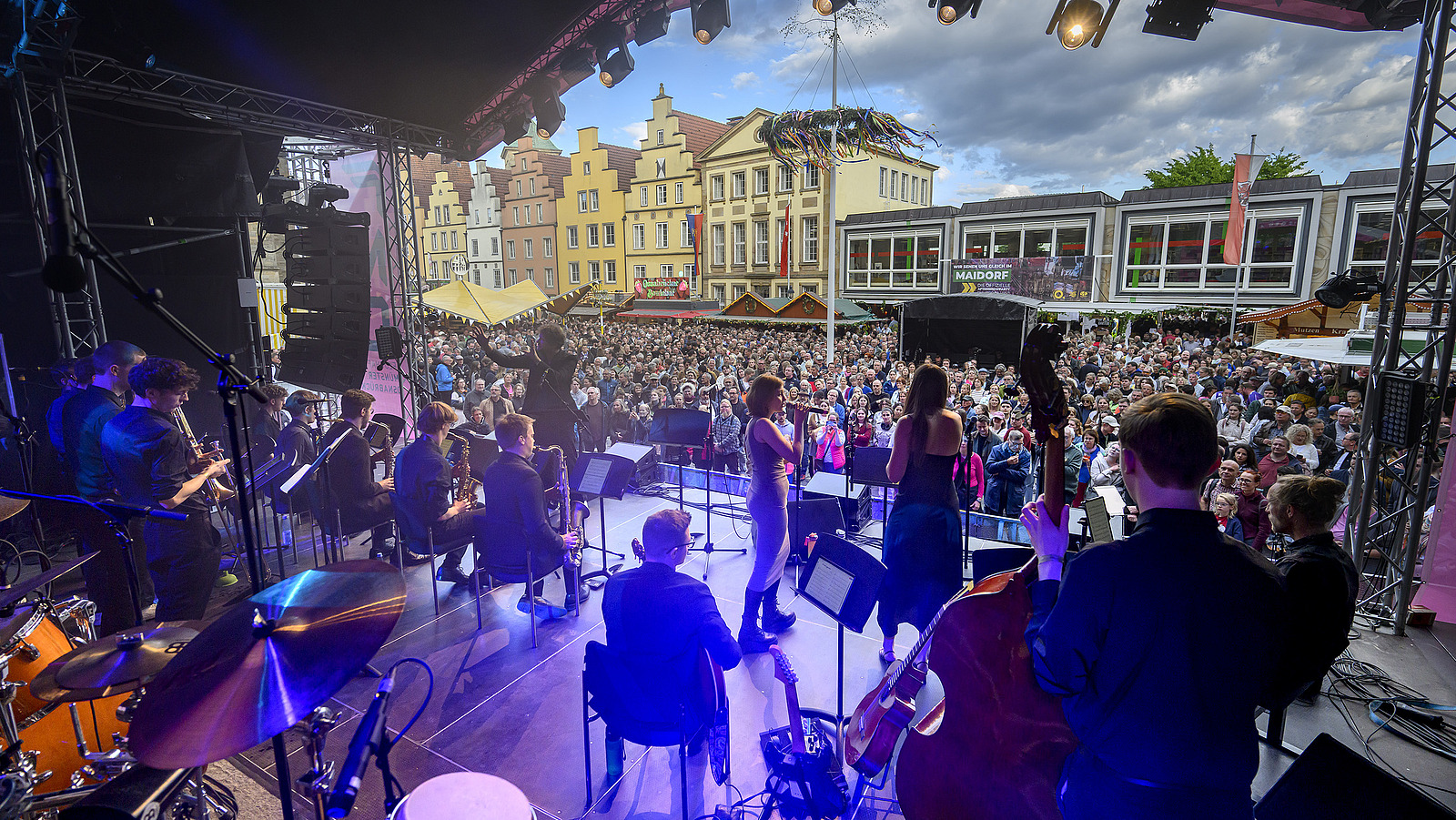 A stage with musicians and many spectators in front of it, with buildings in the background