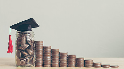 Eine Treppe aus immer größer werdenen Münztürmen führt zu einem Glas voller Münzen, auf dem eine Absolvent*innenkappe liegt. A staircase made of increasingly larger stacks of coins leads to a jar full of coins, on top of which rests a graduation cap.