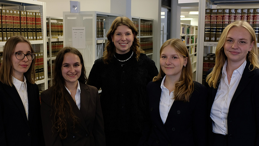 Fünf förmlich gekleidete junge Frauen nebeneinander, im Hintergrund eine Bibliothek