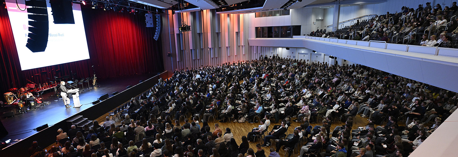 A hall with a seated audience and a person on stage
