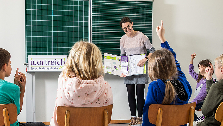 Einige Schüler und Schülerinnen sitzen in einem Stuhlkreis vor einer tafel. Eine Lehrerin hält einen geöffneten wortreich Ordner, einige Schüler melden sich.
