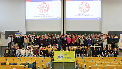 A view of a large group of people standing in a lecture hall and smiling at the camera. In the front row, several people are holding a banner that reads “Young German Physical Society”