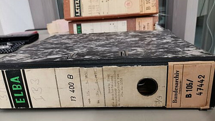 Ein Aktenordner liegt auf einem Tisch. Der Ordner hat ein marmoriertes Muster und trägt Etiketten mit verschiedenen Beschriftungen, darunter "ELBA" und "Bundesarchiv". Im Hintergrund sind weitere Aktenordner gestapelt. Aus einem Ordner ragen Papierbögen heraus. Die Oberfläche des Tisches ist grau, und im Hintergrund sind unscharfe Büroelemente erkennbar.