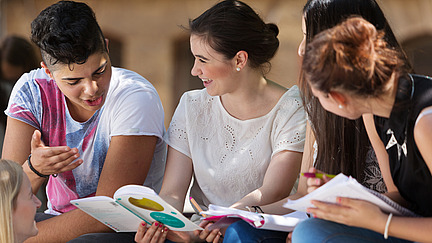 Drei junge Personen sitzen zusammen und schauen fröhlich in ein Buch; Three young people are sitting together and happily looking at a book.