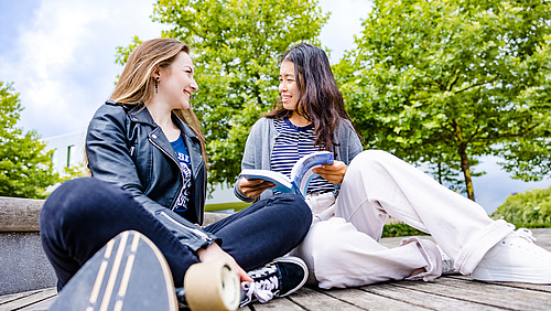 Two female students are sitting outside on campus; one of them is holding a book