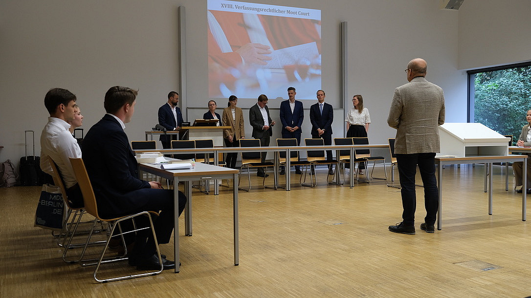 Professor Dörr, im Vordergrund mit dem Rücken zur Kamera stehend, bedankt sich bei den sieben Personen, die hinter einer Reihe von Tischen stehen und die Rolle der Richterinnen und Richter in dieser Simulation gespielt haben. Im Hintergrund ein Bild der roten Roben der Verfassungsrichter. Rechts und links die Teams an Tischen sitzend.