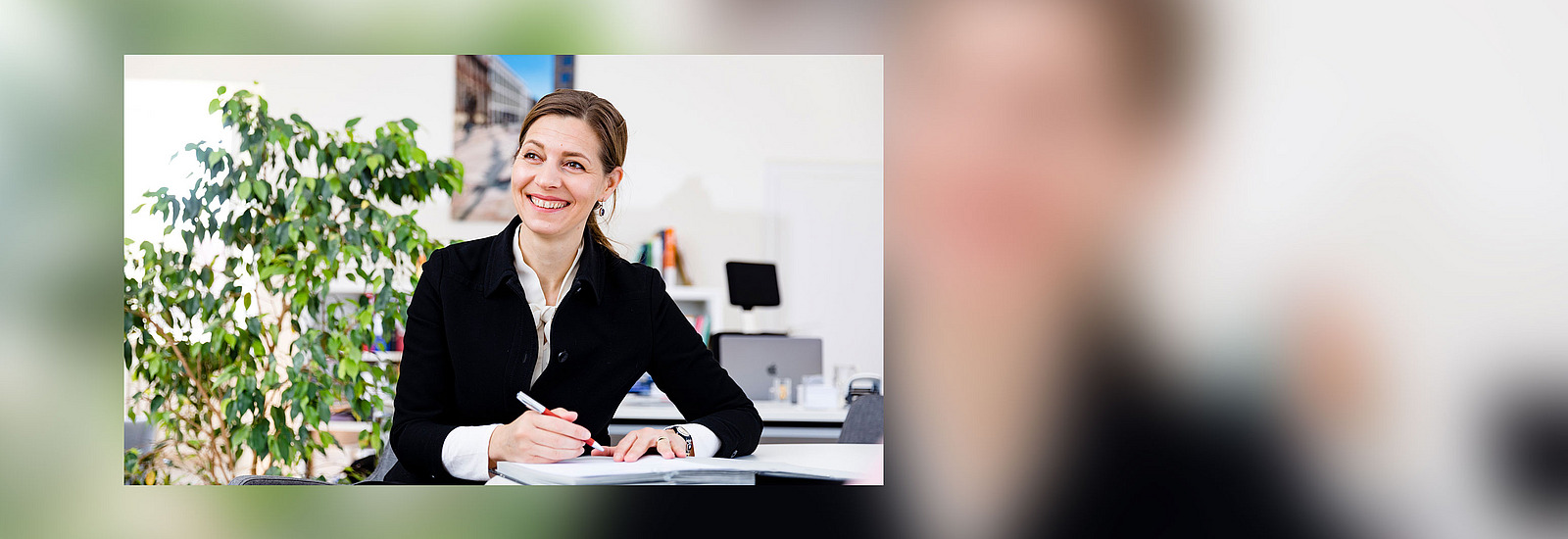 University president Susanne Menzel-Riedl sits at the desk and smiles at someone next to the camera.