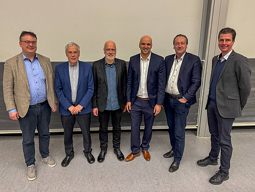 6 M&auml;nner stehen nebeneinander vor einer Tafel und schauen in die Kamera