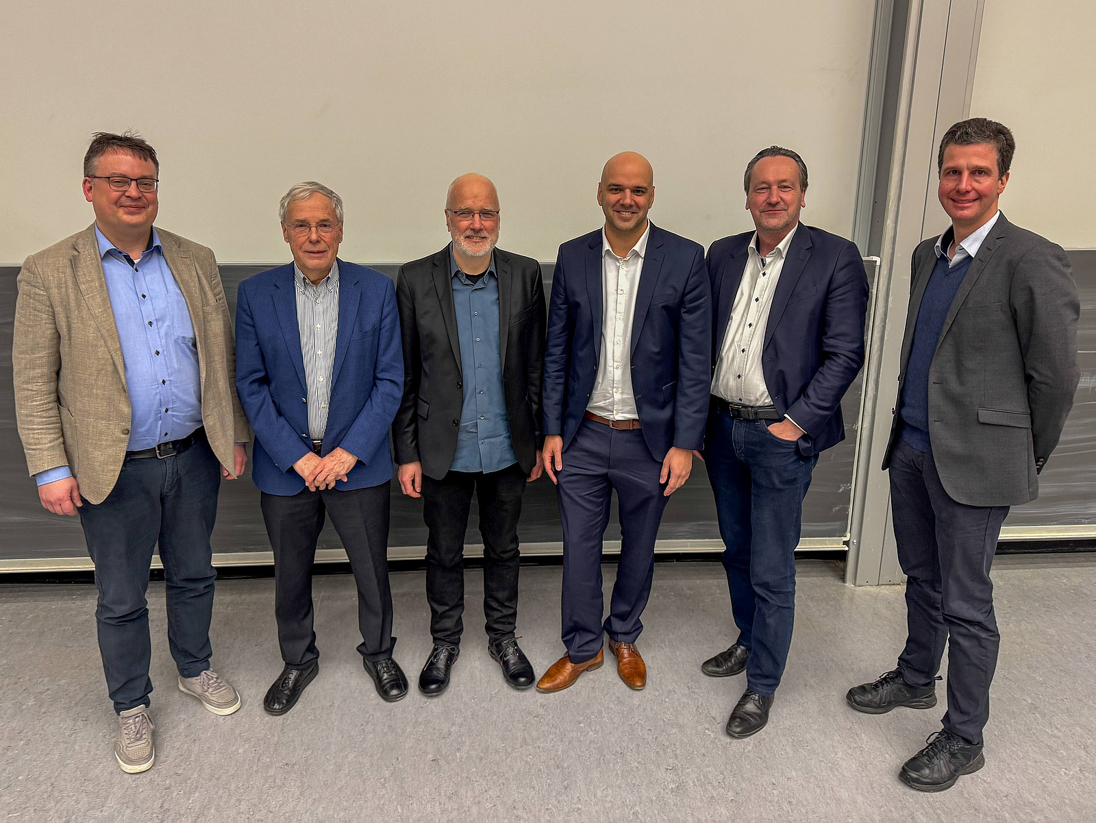 6 M&auml;nner stehen nebeneinander vor einer Tafel und schauen in die Kamera