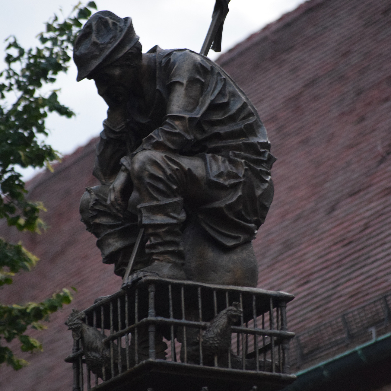 Das Bild zeigt eine Bronzeskulptur. Auf einer Säule sitzt zusammengekauert eine bäuerlich gekleidete Figur.