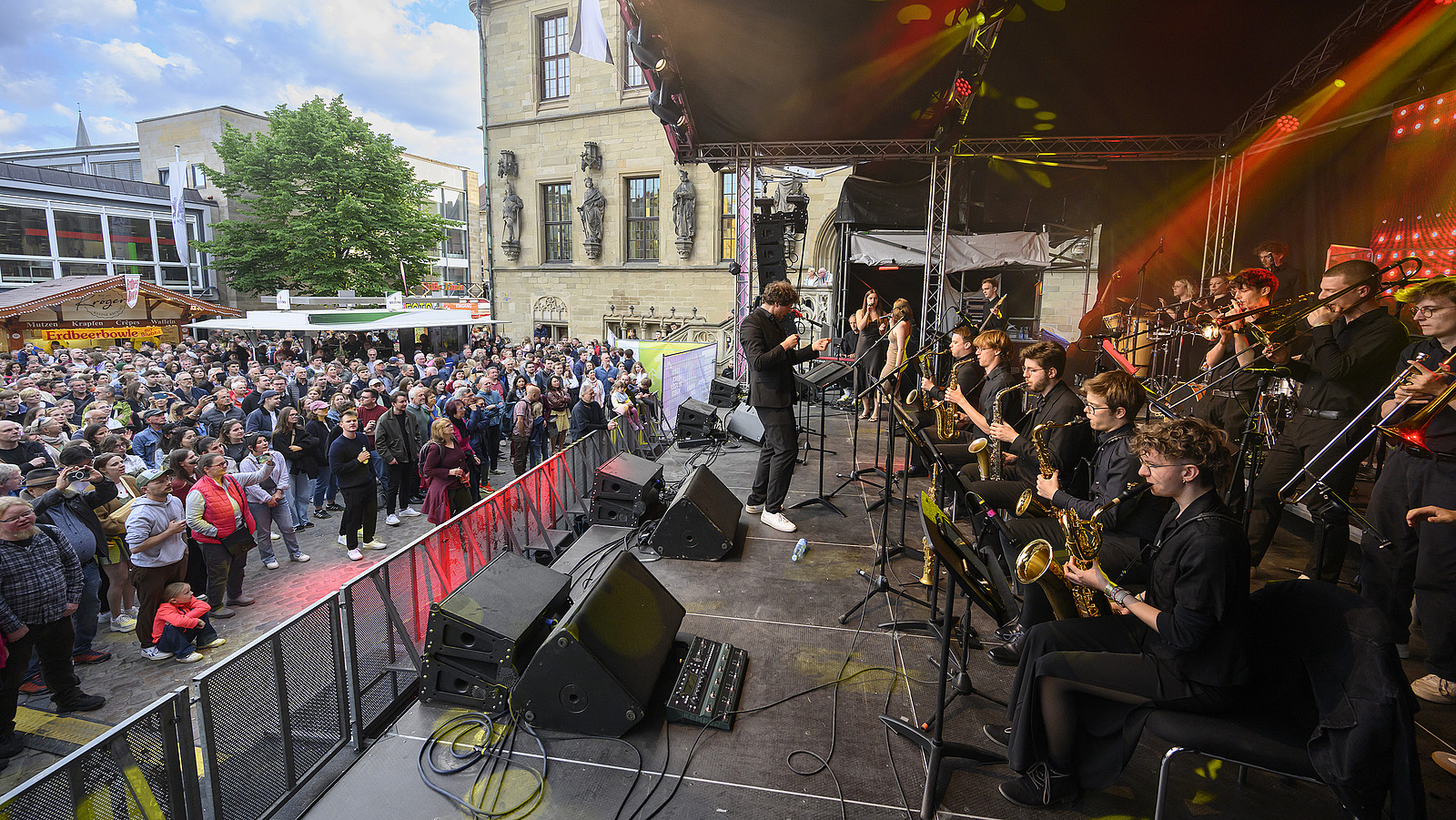 Several brass players on a stage with conductor, many spectators in front of the stage