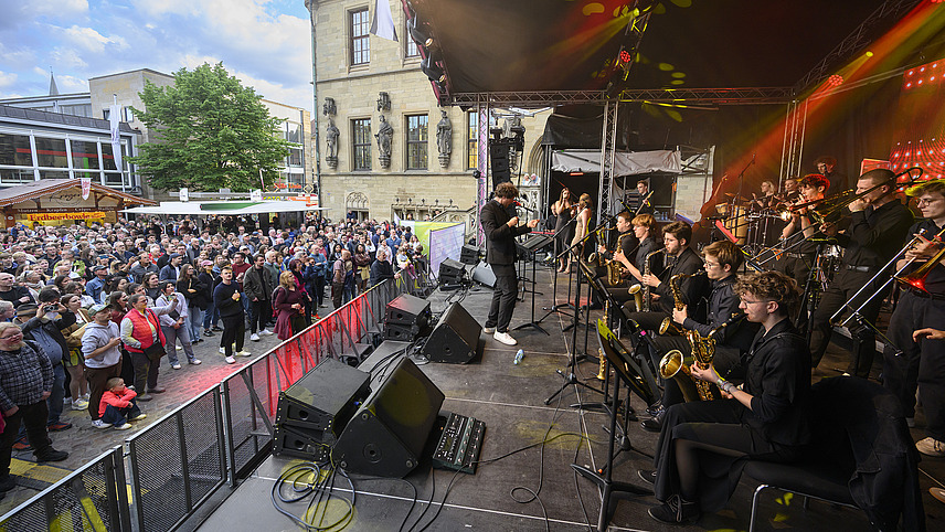 Several brass players on a stage with conductor, many spectators in front of the stage