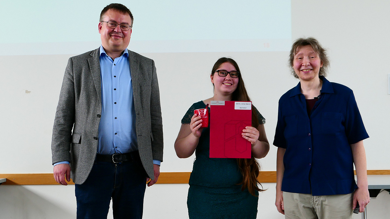 Eine Studentin mit Studiendekanin und Dekan stehen nebeneinander. Die Studentin hat eine Mappe in der Hand.