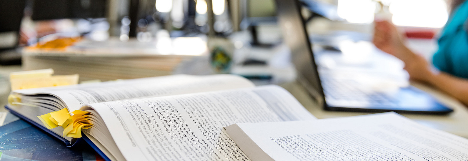 Ein paar offene Bücher an einem Schreibtisch, an dem eine Person arbeitet.