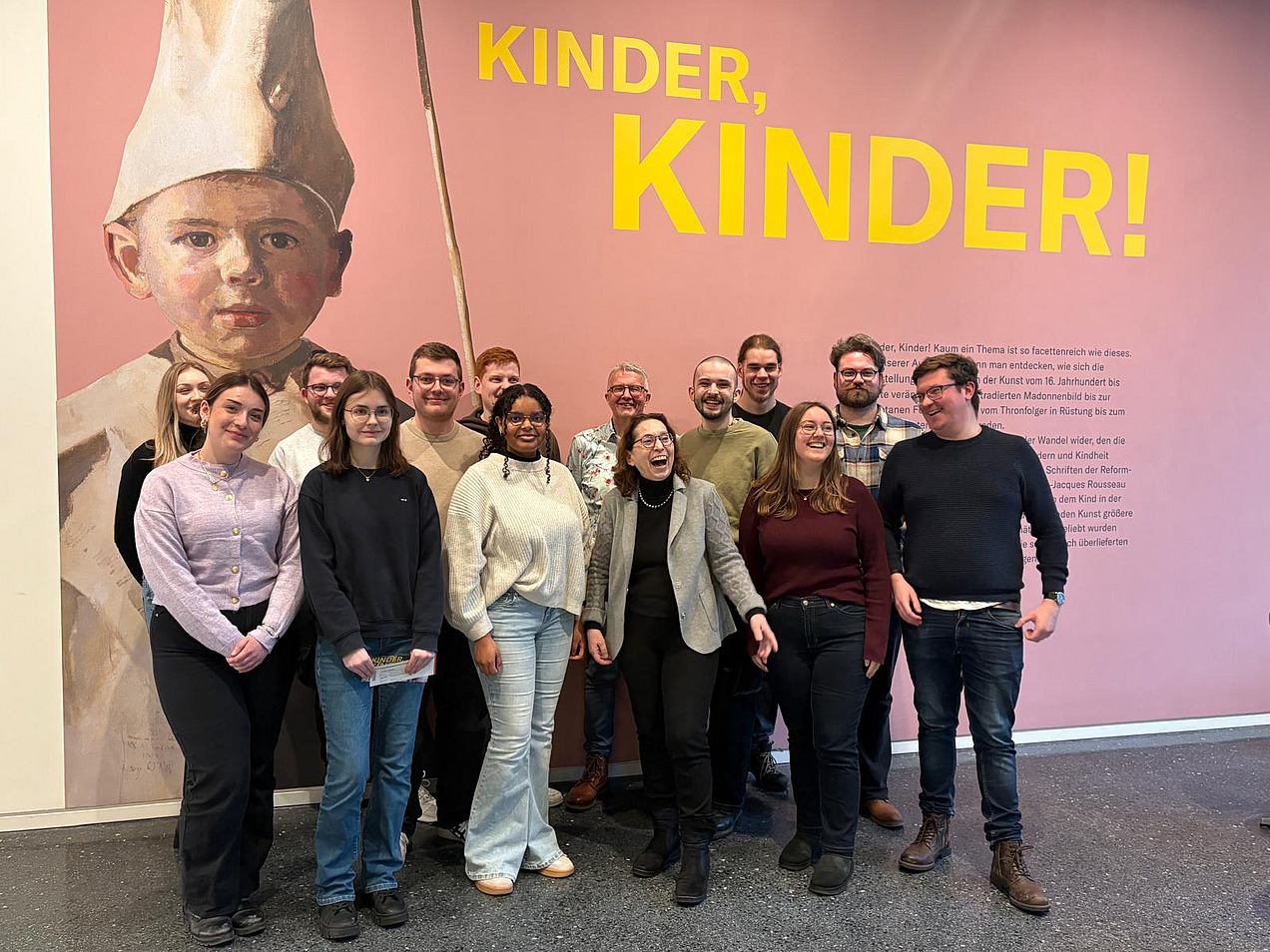 Das Bild zeigt eine Gruppe l&auml;chelnder Menschen vor einer rosafarbenen Wand.