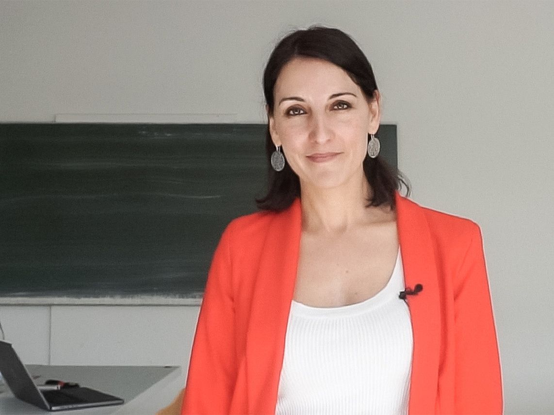 Eine Frau mittleren Alters in Shirt und Jackett steht vor einer Schiefertafel und l&auml;chelt in die Kamera