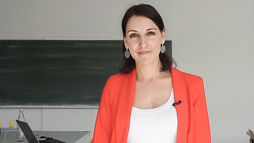 A middle-aged woman in a shirt and jacket stands in front of a slate and smiles at the camera