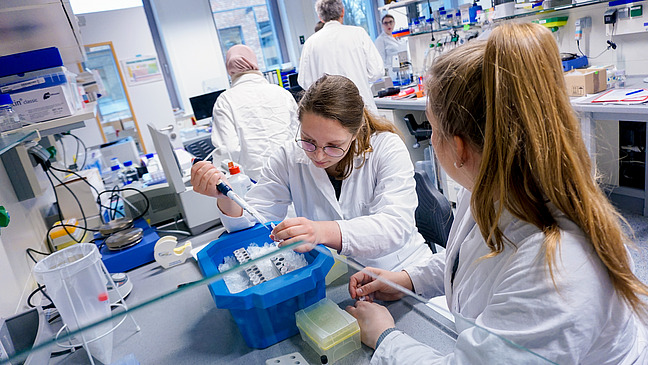 Several people wearing lab coats in a laboratory. A young woman pipettes something into a reaction vessel.