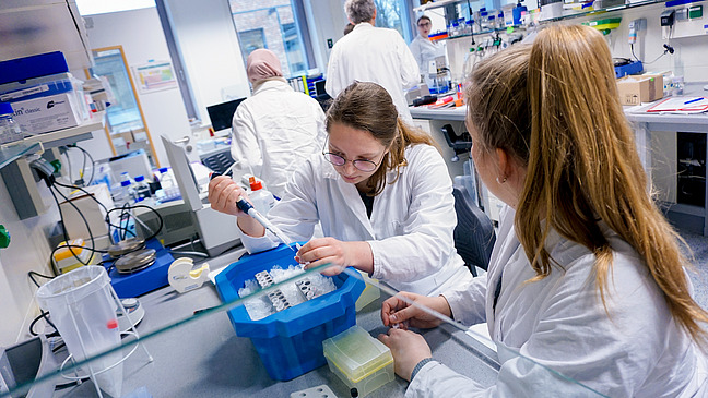Mehrere Personen mit Kitteln in einem Labor. Eine junge Frau pipettiert etwas in ein Reaktionsgef&auml;&szlig;.