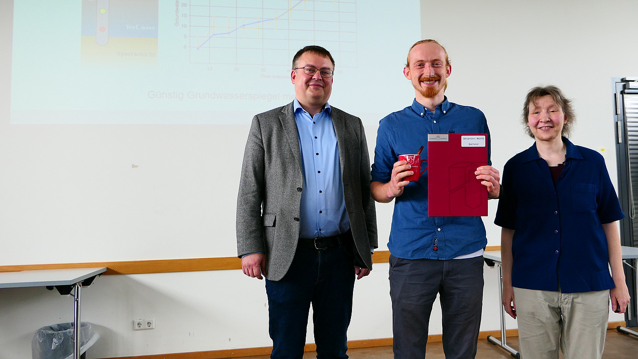 Ein Student mit Studiendekanin und Dekan stehen nebeneinander. Der Student hat eine Mappe in der Hand