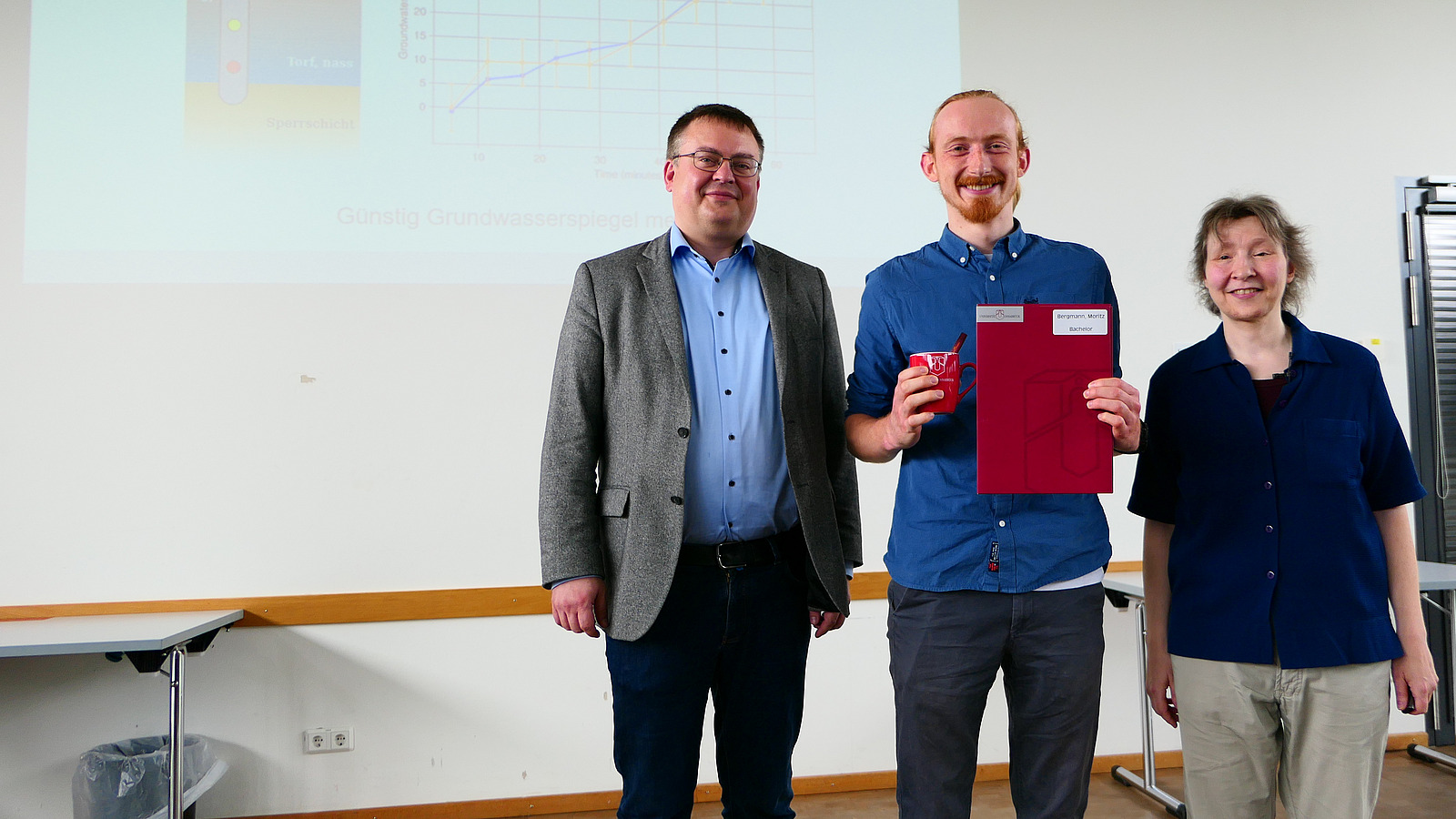 Ein Student mit Studiendekanin und Dekan stehen nebeneinander. Der Student hat eine Mappe in der Hand