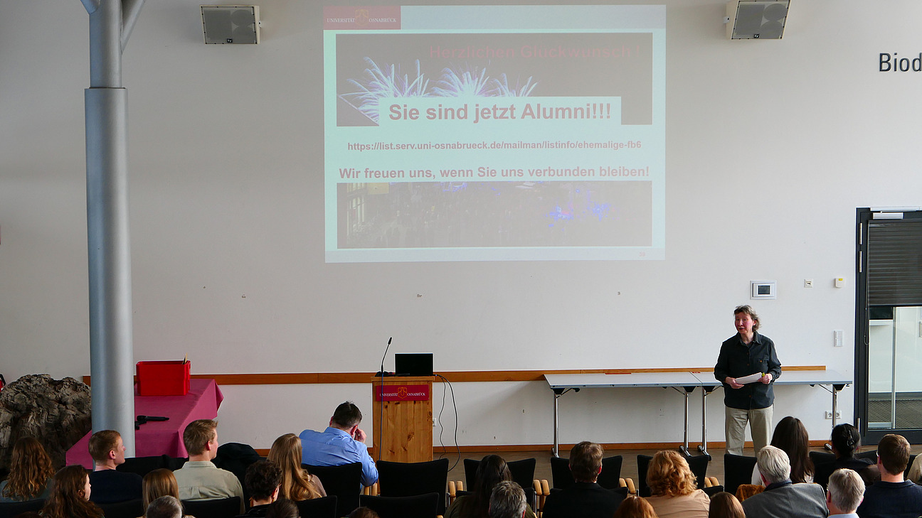 Eine Dozentin steht vor gef&uuml;llten Sitzreihen. Im Hintergrund die Projektion eines Feuerwerks mit Gratulationen.