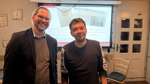 Zwei Personen stehen nebeneinander in einem Raum und schauen in Richtung Kamera.  Hinter ihnen ist eine Projektionsfl&auml;che mit Text und Bildern zu sehen. Au&szlig;erdem stehen im Raum mehrere St&uuml;hle.  Die beiden Personen stehen im Vordergrund und nehmen den gr&ouml;&szlig;ten Teil des Bildes ein.