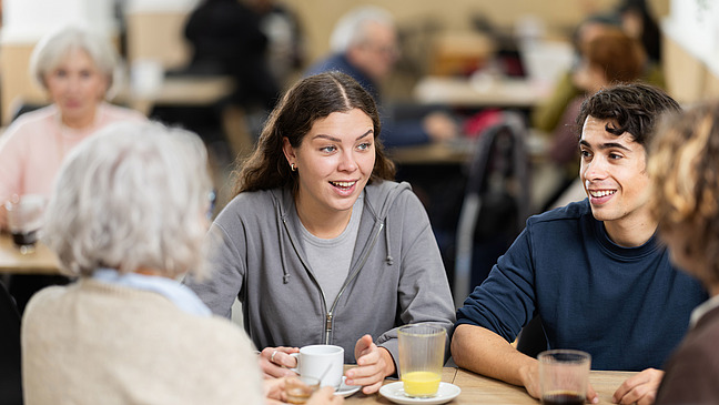 Mehrere Personen verschiedener Altersstufen sitzen mit Getr&auml;nken an einem Tisch und unterhalten sich