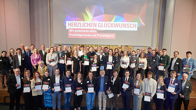 Eine Gruppe von Menschen steht auf einer B&uuml;hne und h&auml;lt Urkunden in den H&auml;nden. Die Personen l&auml;cheln und sind in formeller Kleidung.