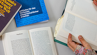 Ausgebreitete B&uuml;cher auf einem Tisch, dazwischen H&auml;nde von Studierenden, die sich Notizen in aufgeklappten Heften aufschreiben.